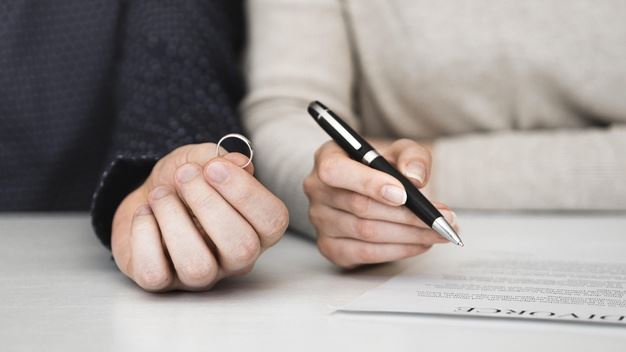 One hand holding a ring, another hand holding a pen poised above a divorce document.